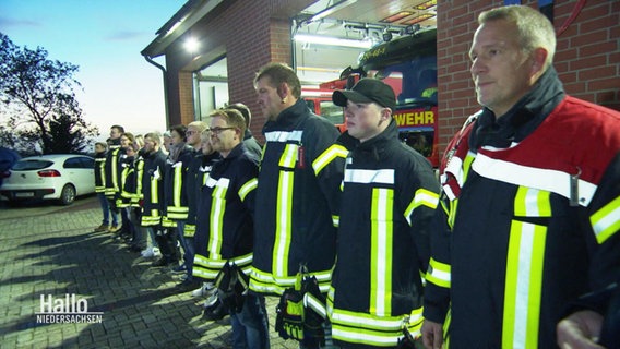 Feuerwehrleute stehen in einer Reihe vor ihrer Feuerwehrwache und schauen geradeaus. © Screenshot 