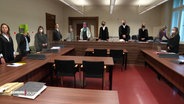 Mehrere Menschen stehen in einem Gerichtssaal, eine Frau hält sich eine Mappe vors Gesicht. © Screenshot 