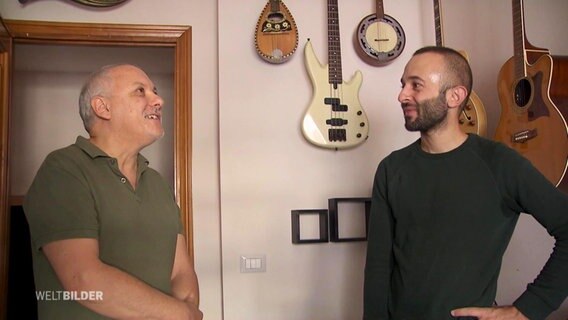 Zwei Männer stehen vor einer Wand, an der Gitarren hängen. © Screenshot 