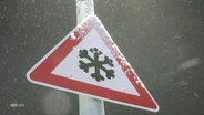 Schneegestöber. An einem Pfosten ein dreieckiges Warnschild mit einem Eiskristall in der Mitte. © Screenshot 