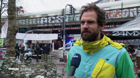 Ein junger Mann im Interview, im Hintergrund eine Zugbrücke und protestierende Menschen. © Screenshot 