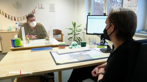Zwei Menschen sitzen in einem Büro an Tischen und sprechen miteinander. © Screenshot 