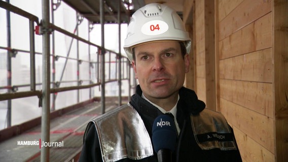 Ein Mann mit Schutzhelm im Interview, im Hintergrund eine Holzfassade. © Screenshot 