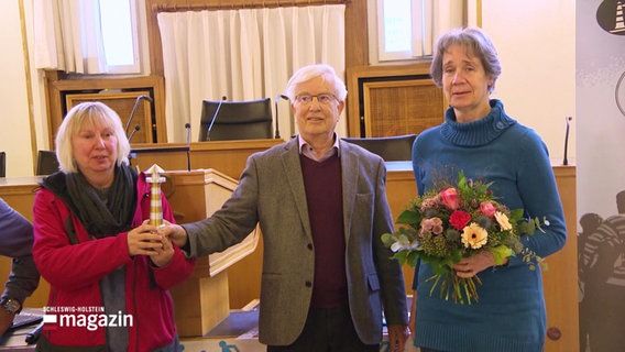 Drei Menschen bei der Preisverleihung, sie halten eine kleinen Leuchtturm und einen Blumenstrauss. © Screenshot 