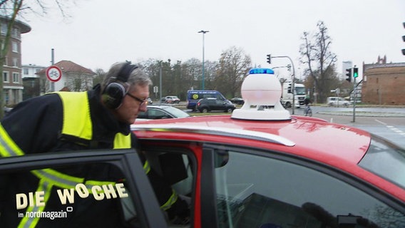 Ein Mann mit Kopfhörern und Signalweste neben einem Auto, das eine Sirene auf dem Dach installiert hat. © Screenshot 