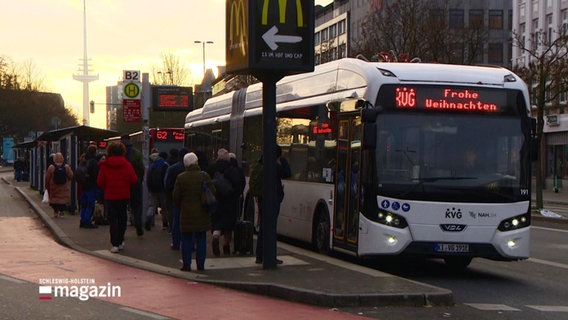 Szene an einem Busbahnhof: Wartende Menschen, zwei Busse halten, auf einer Busanzeige steht: "KVG - Frohe Weihnachten". © Screenshot 