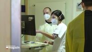 Eine Mitarbeiterin und ein Mitarbeiter des medizinischen Personals in einer Klinik an einem Bildschirm, beide tragen Schutzkleidung. © Screenshot 