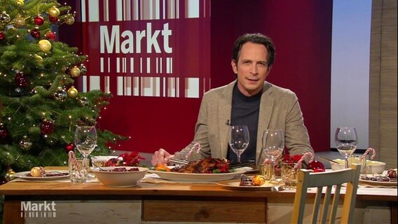 Im Studio von "Markt": Moderator Jo Hiller sitzt an einem weihnachtlich geschmüchten und gedeckten Tisch, vor sich einen Braten. Links im Anschinitt steht ein geschmückter Weihnachtsbaum. © Screenshot 