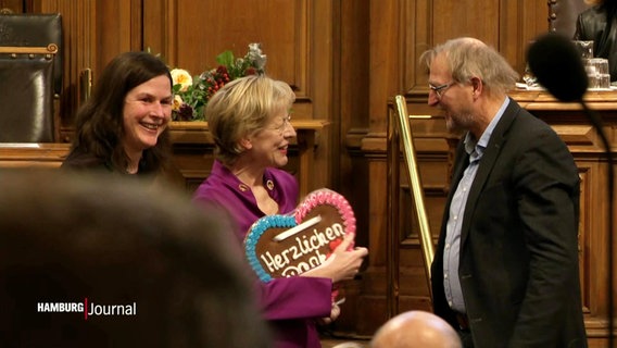 Dorothee Stapelfeld wird mit Lebkuchenherz in der Hamburgischen Bürgerschaft verabschiedet. Stapelfeld lacht einen Kollegen an und trägt ein violettes Kostüm. © Screenshot 