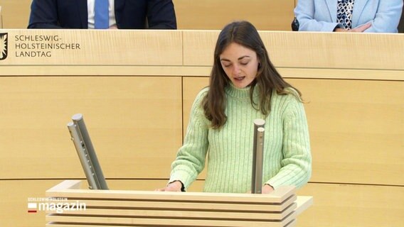 Nelly Waldeck von den Grünen steht am Rednerpult. Sie hat lange braune Haare und trägt einen hellgrünen Pullover. © Screenshot 