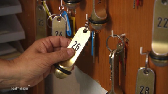 Eine Hand hebt einen Schlüsselbund zu einem Gästezimmer von einem Haken an einem Schlüsselbrett ab. © Screenshot 