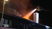 Ein Personal der Feuerwehr löscht einen Großbrand an einer Lagerhalle bei Nacht vom Drehleiterkorb aus. © Screenshot 