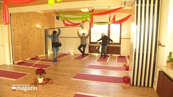 Eine Frau und ein Mann machen Yoga-Übungen auf Gymnastikmatten in einem Innenraum. Ein Tonmann steht daneben und hält eine Tonangel. © Screenshot 
