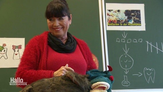 Die Zahnfee, Christiane Lotties, steht vor einer Tafel in einem Klassenzimmer und lächelt. © Screenshot 