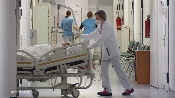 Eine Frau in weißer Arbeitskleidung schiebt auf einem Gang eines Krankenhauses ein Krankenbett in einen Seitengang. © Screenshot 