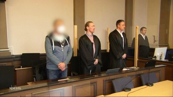Der Angeklagte und seine Verteidigung stehen im Gerichtssaal © Screenshot 