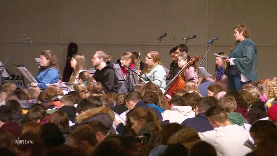 Viele Jugendliche sitzen zusammen in einem großen Raum, im Hintergrund musizieren welche. © Screenshot 