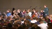 Viele Jugendliche sitzen zusammen in einem großen Raum, im Hintergrund musizieren welche. © Screenshot 