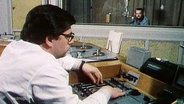 Eine Person sitzt vor einem Tonbandgerät, im Hintergrund sitzt jemand in einem Studio © Screenshot 