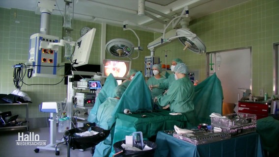 Krankenhauspersonal steht eingekleidet in hyginische Schutzausrüstung an einem OP-Tisch im Operationssaal. © Screenshot 