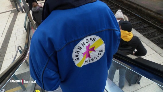 Eine Person mit dem Emblem der Bahnhosmission auf der Jacke fährt mit einer Rolltreppe. © Screenshot 