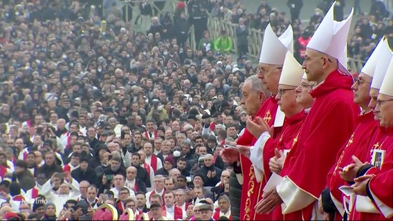 Vor einer riesigen Menschenmenge stehen mehrere katholische Priester und Geistliche in rotren Gewändern mit Mitra-Kappen im Profil zu Kamera bei einer Gedenkveranstaltung. © Screenshot 