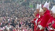Vor einer riesigen Menschenmenge stehen mehrere katholische Priester und Geistliche in rotren Gewändern mit Mitra-Kappen im Profil zu Kamera bei einer Gedenkveranstaltung. © Screenshot 