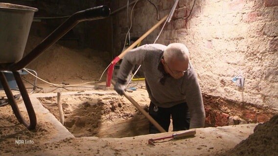 Ein Mann steht bis zur Hüfte in einem Loch in einem gemauerten Raum und schaufelt Erde. © Screenshot 