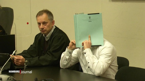 Der Angeklagte mit einer Mappe vor dem Gesicht im Gerichtssaal. Neben ihm ein Jurist. © Screenshot 