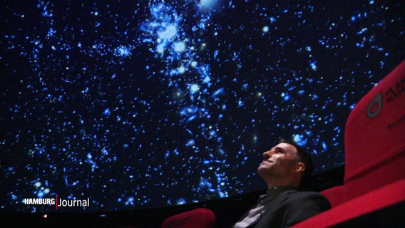Ein Mann sitzt in einem roten Kinosessel und schaut auf die Leinwand unter der Decke des Planetariums. © Screenshot 