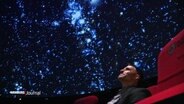 Ein Mann sitzt in einem roten Kinosessel und schaut auf die Leinwand unter der Decke des Planetariums. © Screenshot 