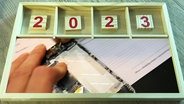 Ein Holzkasten, in dem die Zahlen "2023" liegen. Im unteren Segment ist ein Bild einer Hand mit einem kaputten Handy zu sehen. © Screenshot 