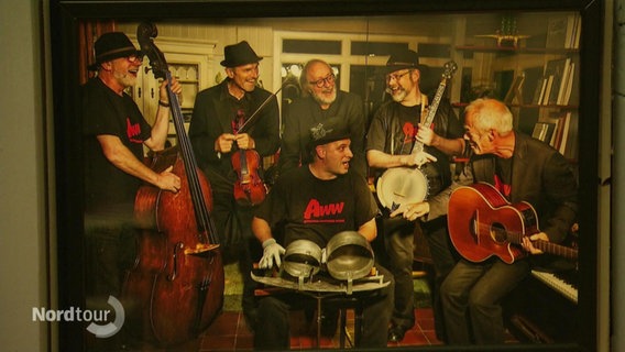 Auf einem eingerahmten Bild an einer Wand ist eine Gruppe älterer Musiker zu sehen die sich zusammen lachen und typische Folk-Instrumente bei sich haben. © Screenshot 