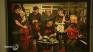 Auf einem eingerahmten Bild an einer Wand ist eine Gruppe älterer Musiker zu sehen die sich zusammen lachen und typische Folk-Instrumente bei sich haben. © Screenshot 
