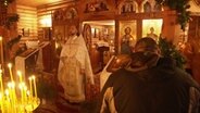 Im Altarraum einer kleinen russisch-orthodoxen Kirche steht beim Weihnachtsgottesdienst ein Erzpriester in einer feierlicher Robe vor der Gemeinde. © Screenshot 