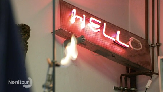 Eine Leuchtstoffröhre in Form des Wortes Hello hängt an einer Wand. Darunter eine Flamme. © Screenshot 