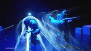 Eine Tanzperformance auf einer Theaterbühne, bei der die Tanzenden von verschieden farbigen Lichtern angestrahlt werden. © Screenshot 