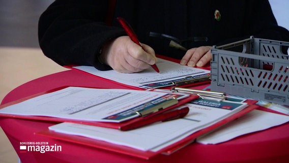 Eine Person schreibt auf einem roten Klemmbrett. © Screenshot 