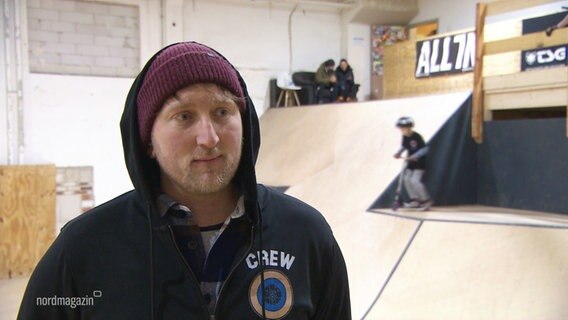 Ein Mann mit einer Kapuze über einer Wollmütze in einer Skater-Halle im Interview. © Screenshot 