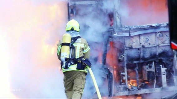 Eine Einsatzkraft der Feuerwehr steht am ausgebrannten Wrack eines Linienbusses und löscht letzte Flammen. © Screenshot 