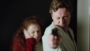 Eine Person mit Waffe, die zielt. Eine Person im Hintergund die sich die Ohren zuhält © Screenshot 