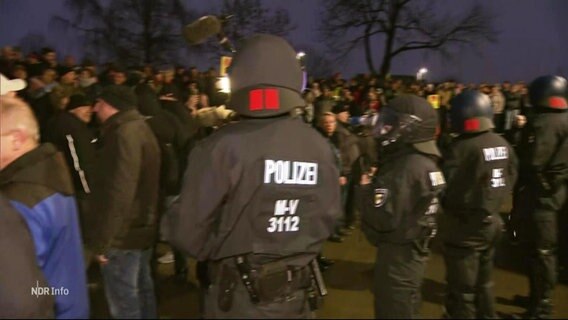 Vor dem Kreistagsgebäude hat sich eine Reihe von Einsatzkräften der Polizei in Demonstrationsschutzausrüstung aufgestellt, um eine wütende Gruppe von Menschen abzuhalten. © Screenshot 