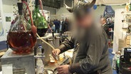 In einem Messestand füllt ein unkenntlich gemachter Mann in einem Wikingerkostüm ein Getränk an einer Zapfsäule in ein Glas. © Screenshot 