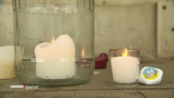 Auf einem gepflasterten Fußboden stehen mehrere Gedenkkerzen sowie ein kleiner Stein mit einer Friedenstaube darauf. © Screenshot 