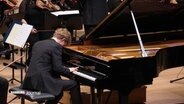 Pianist Jan Liesiecki spielt am Flügel auf der Bühne vor dem Royal Philharmonic Orchestra im großen Saal der Elbphilharmonie. © Screenshot 