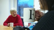 Im Vordergrund: Eine Frau trinkt aus einer Tasse mit dem Buchstaben E darauf. Im Hintergrund: Ihr gegenüber am Tisch sitzt eine Frau, die ihr zulächelt. © Screenshot 