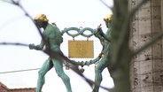 Zwei Figuren aus Bronze an einer Häuserwand, sie tragen zwischen sich einen vergoldeten Keks. © Screenshot 