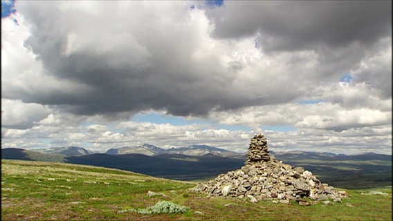 Ein Steinhügel auf einem Rasen vor Mittelgebirgspanorama mit Wolken am Himmel. © Screenshot 