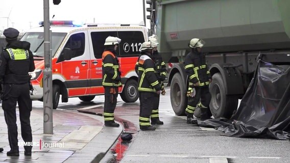 Einsatzkräfte von Feuerwehr und Polizei sichern eine Unfallstelle ab, an der eine Radfahrerin von einem Lkw überrollt wurde. Der Bereiche unter dem Lkw ist mit einer schwarzen Plastplane verhängt. © Screenshot 