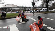 An einem Ampelübergang an der Hamburger Kennedybrücke haben sich Protestierende mit Warnwesten und Transparenten auf die Fahrbahn gesetzt. © Screenshot 